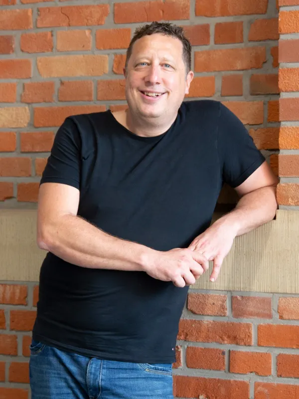 Steffen Jung lächelnd vor einer Backsteinwand, in schwarzem T-Shirt